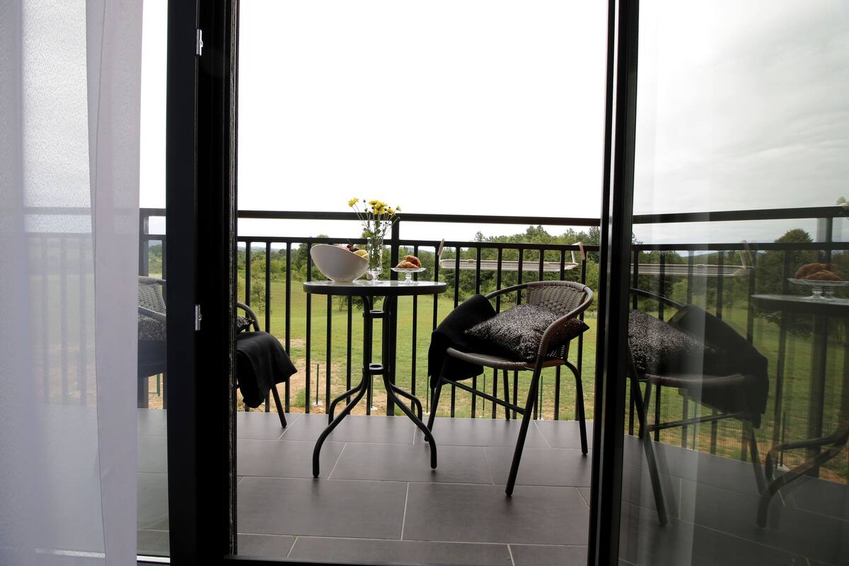 Un balcón con una mesa y sillas, un florero con flores, y vistas a un paisaje verde.