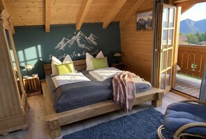 Ein Schlafzimmer in einer Holzhütte mit einem Bett, Kissen, einem Nachttisch, einer Lampe und Blick auf Berge durch eine Tür.