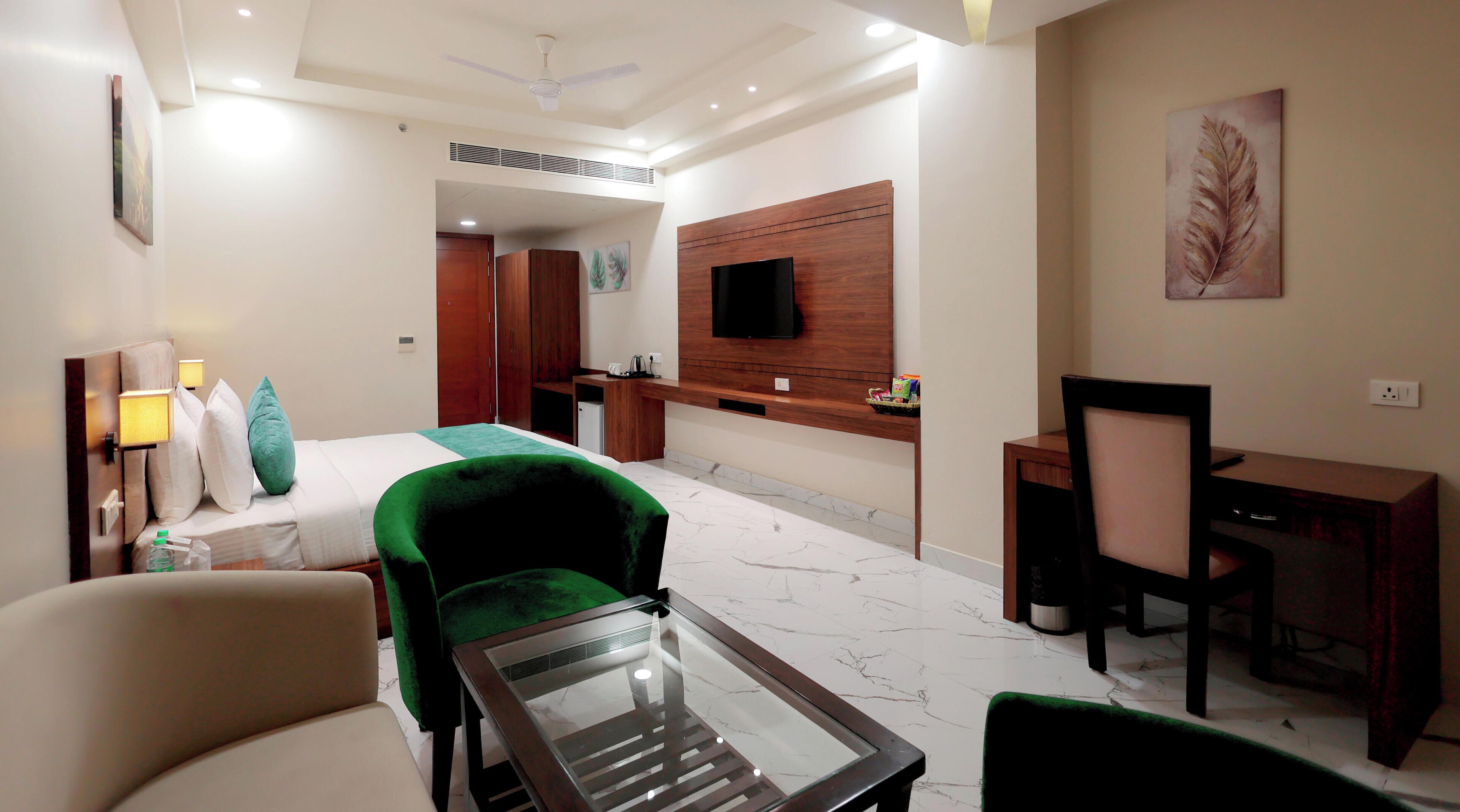 Habitación de hotel con cama, una silla verde, escritorio, televisor montado en la pared y un baño visible a través de una puerta abierta.