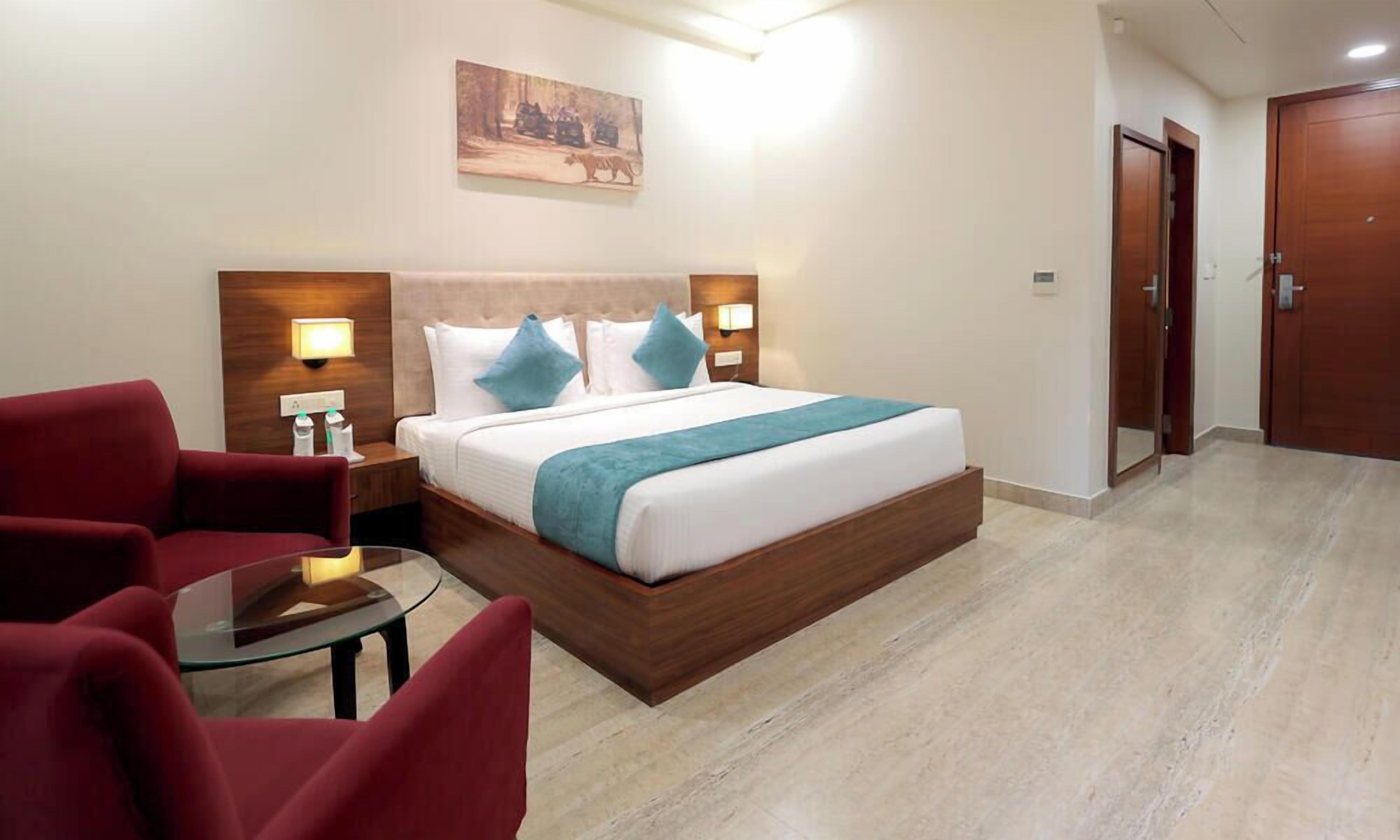 Habitación de hotel con cama, dos mesitas de noche, un sillón rojo, una mesa de vidrio y un cuadro en la pared.
