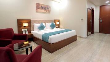 Habitación de hotel con cama, dos mesitas de noche, un sillón rojo, una mesa de vidrio y un cuadro en la pared.