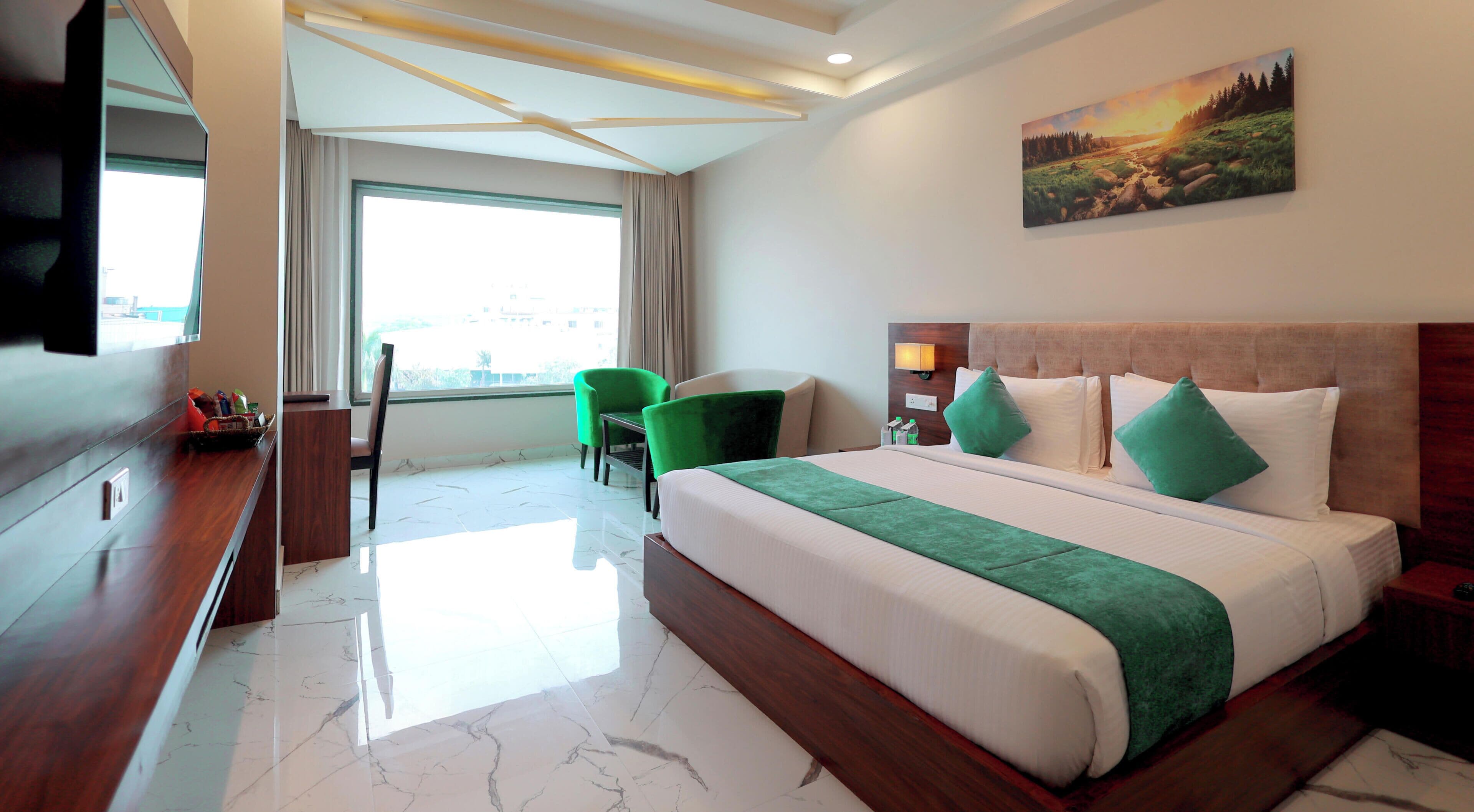 Una habitación de hotel con cama, sillas verdes, televisor, un cuadro y una ventana.