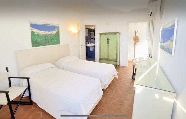 A bedroom with two beds, a green wardrobe, a bathroom with a sink, and a painting of a coastal scene.