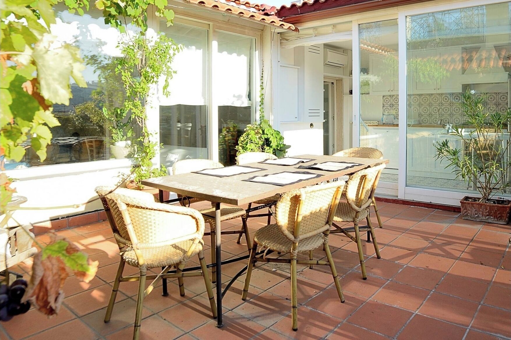 Eine überdachte Terrasse mit Tisch und Stühlen, umgeben von Topfpflanzen und einer Glastür, die in einen Garten führt.