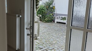 Une porte ouverte donne sur une cour intérieure où l’on aperçoit un radiateur blanc, un banc et une petite plante.