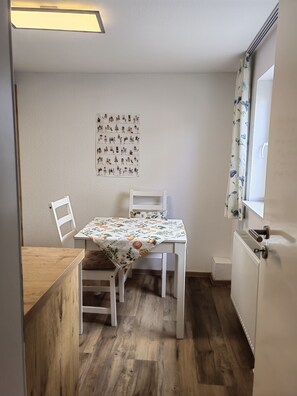 A small, tidy room with a wooden table and two white chairs, a radiator, and a window with floral curtains.