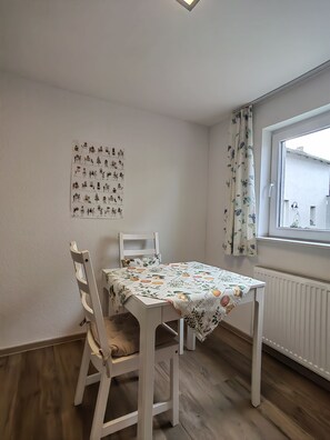 Un coin repas comprenant une table et des chaises, une fenêtre avec des rideaux et un mur orné d’une œuvre d’art à motifs.