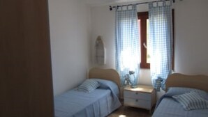 A small, simple bedroom with two single beds, a nightstand, and a window with checkered curtains.