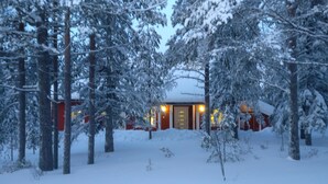 Una cabaña cubierta de nieve con una puerta de madera central y fachada roja, rodeada de árboles cargados de nieve.
