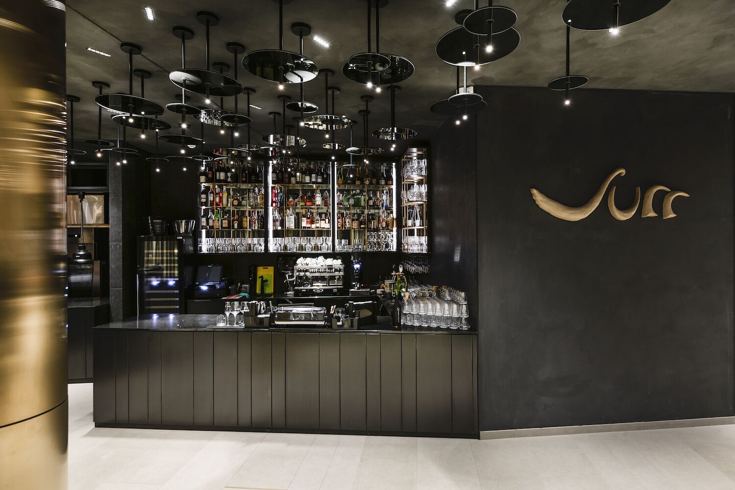 Zona de bar moderna con mostrador negro, varias lámparas colgantes y una variedad de botellas y vasos en estantes.