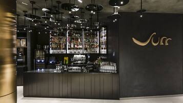 Zona de bar moderna con mostrador negro, varias lámparas colgantes y una variedad de botellas y vasos en estantes.