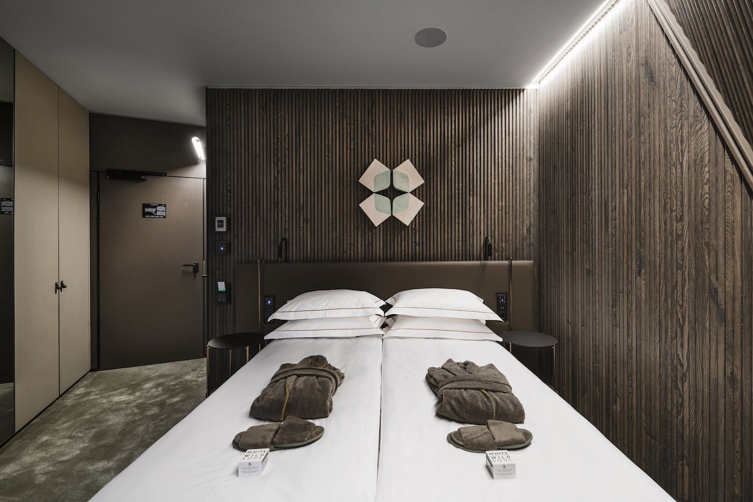 Ein Hotelzimmer mit Bett, Nachttisch, Wandlampe und Wandverkleidung aus Holz.