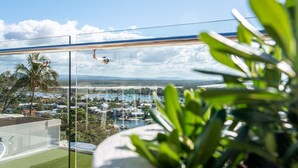 Ein Balkon mit Glasgeländer, der auf eine Küstenstadt blickt, unter wolkenlosem Himmel, mit etwas Grün im Vordergrund.