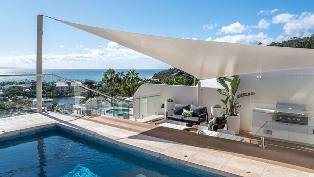 Ein moderner Poolbereich im Freien mit Blick auf den Ozean, einem Grillplatz und einer Lounge-Ecke mit Kissen und Pflanzen.