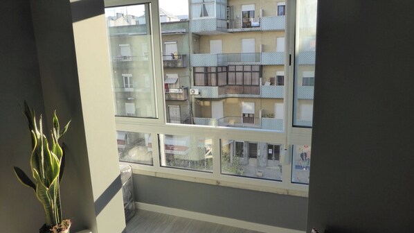 Un balcon donnant sur un immeuble à plusieurs étages, une plante en pot et une fenêtre avec des stores.