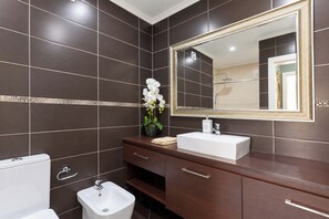 A modern bathroom with dark tiles, a large mirror, a white sink, and a bidet.