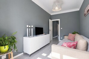 Una sala de estar moderna con una pared gris, un mueble blanco para entretenimiento, un televisor de pantalla plana, un sofá beige con cojines estampados, una planta en maceta verde y una lámpara colgante.