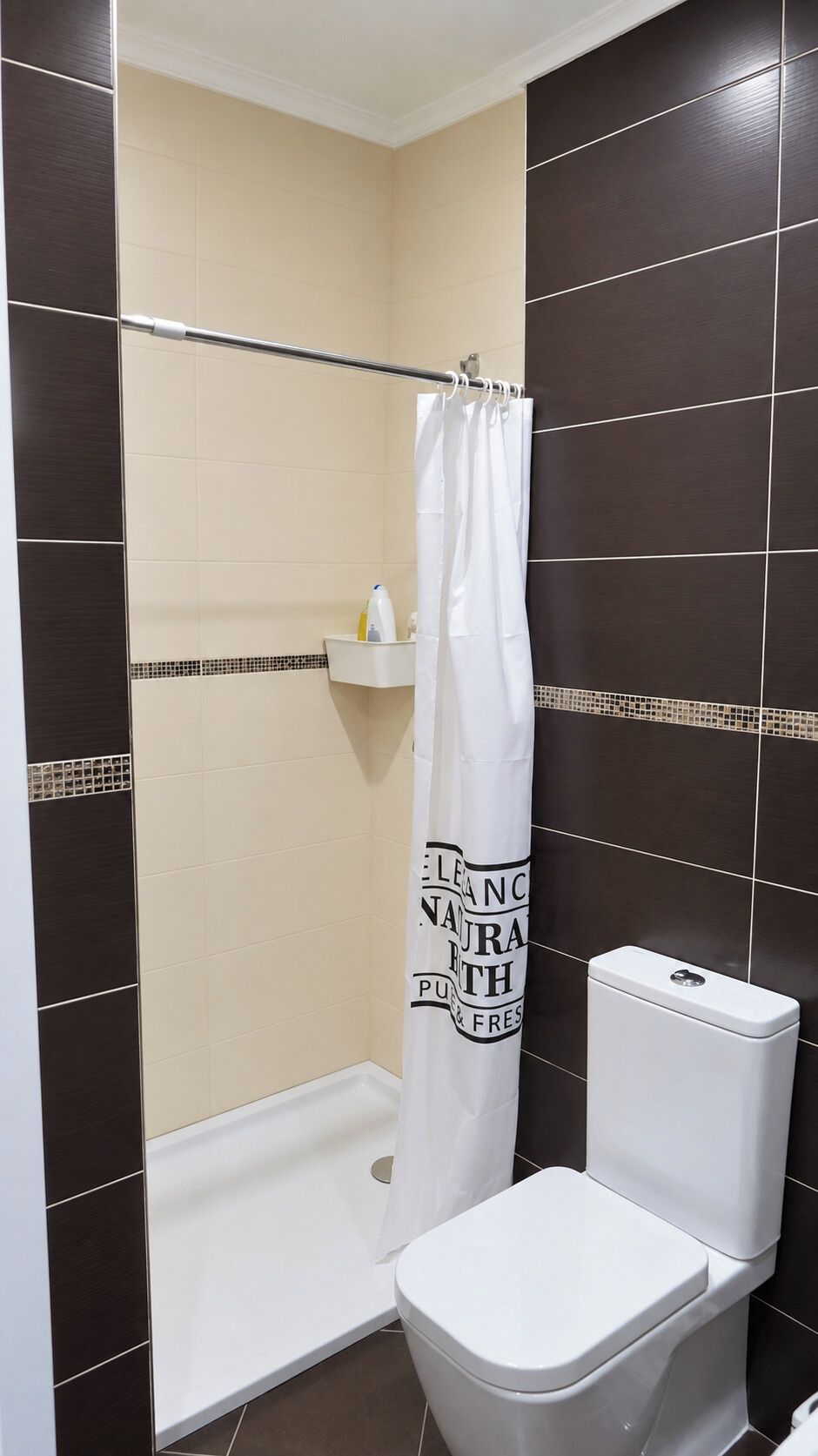 Une salle de bain avec des toilettes blanches, un rideau de douche et des murs recouverts de carreaux de couleur marron foncé.