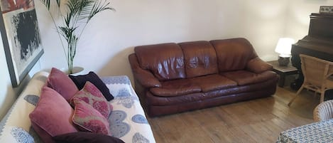 A living room with a brown leather sofa, a patterned sofa, a small table with a lamp, a wicker chair, and a potted plant.