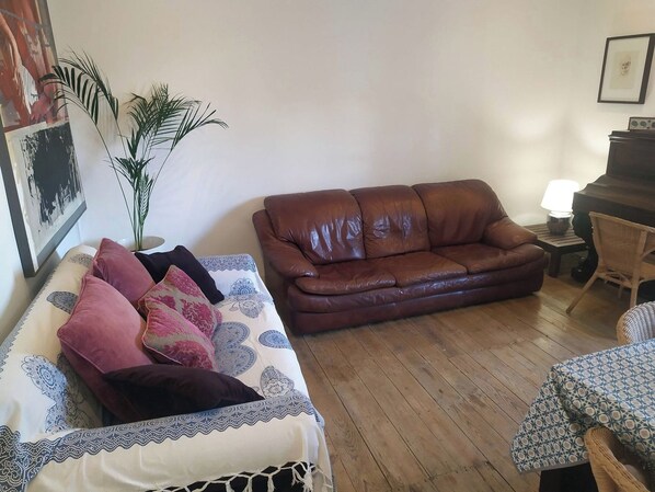 A living room with a brown leather sofa, a patterned sofa, a small table with a lamp, a wicker chair, and a potted plant.