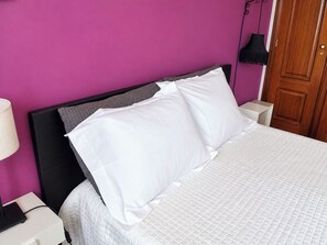 Een bed met witte beddengoed en kussens, een zwart hoofdeinde, een nachtkastje met een lamp, en een houten deur.