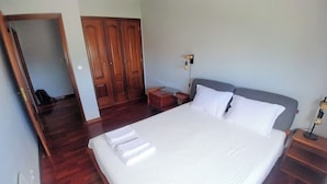 Una cama bien hecha con sábanas y almohadas blancas, un armario de madera, una mesita de noche con lámpara y un aplique de pared.