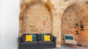Una stanza con parete in pietra, un divano grigio scuro con cuscini gialli e decorati, una sedia bianca e un tappeto verde.