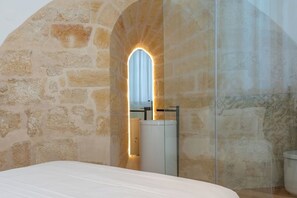 A bedroom with a stone wall, a bed, and a modern bathroom with a sink and mirror. - Historic house of Santo André • Lisbon's old town (Lisboa)