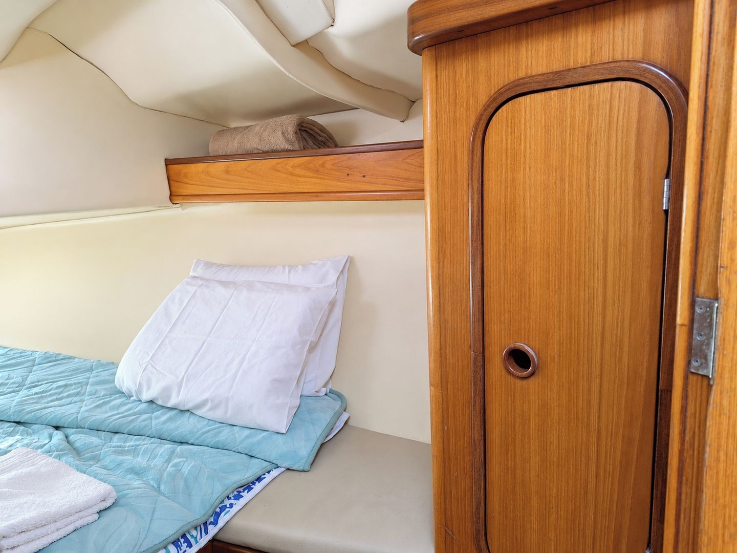Un letto con copriletto azzurro trapuntato e un cuscino bianco, un armadio in legno con maniglia rotonda e uno scaffale sopra il letto.