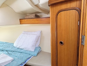 Un lit recouvert d’une couverture matelassée bleue et surmonté d’un oreiller blanc, une armoire en bois dotée d’une poignée ronde, et une étagère au-dessus du lit.
