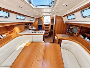 Interior de barco con una mesa de comedor central, asientos, una cocina pequeña y un panel de control.