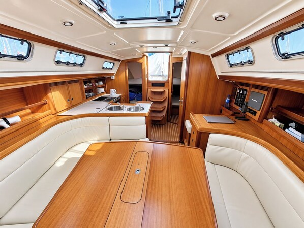 Interior de barco con una mesa de comedor central, asientos, una cocina pequeña y un panel de control.