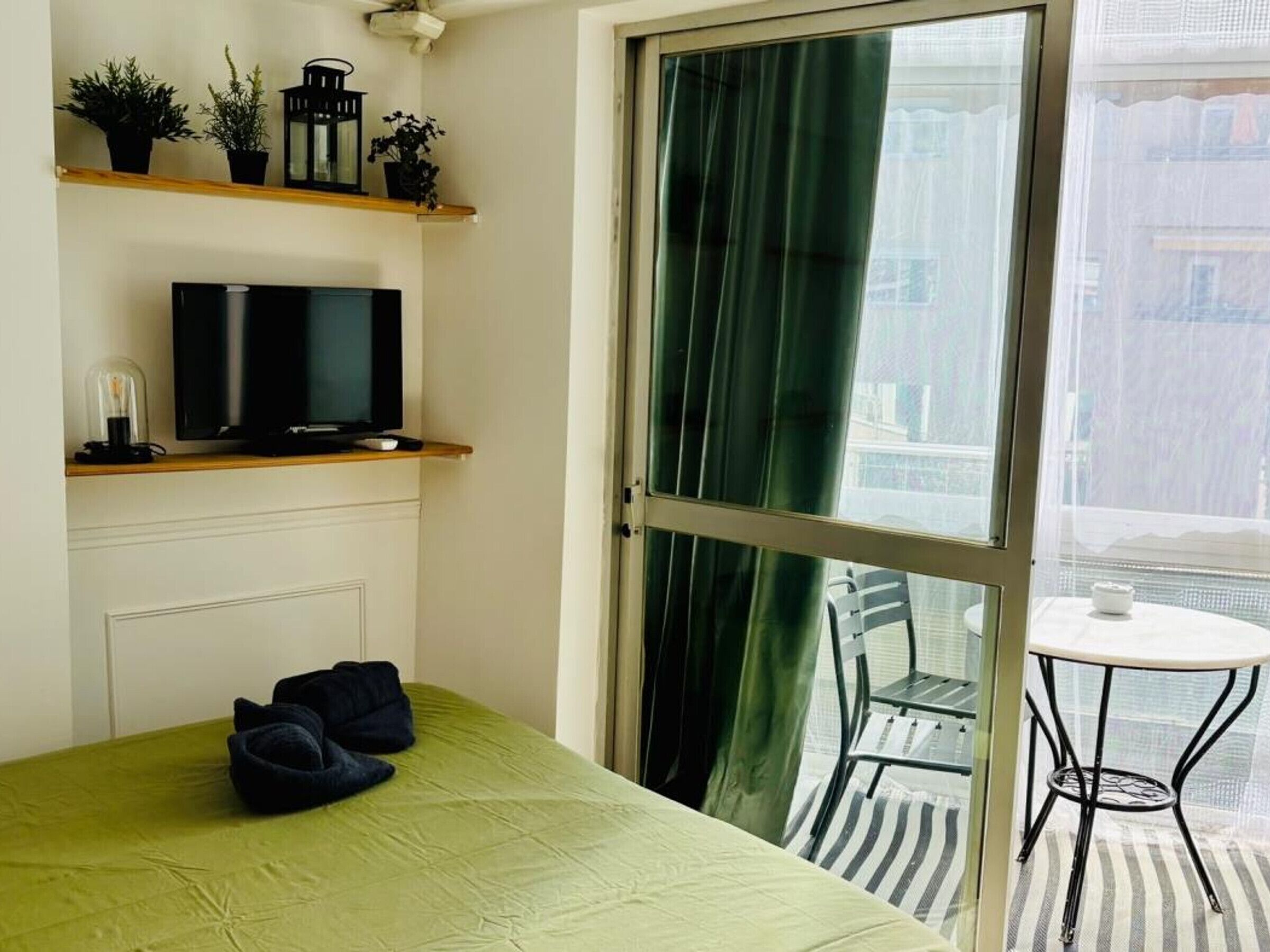 Ein Zimmer mit Bett, Fernseher auf einem Regal, einem Balkon mit Tisch und Stühlen sowie Blick auf Gebäude.