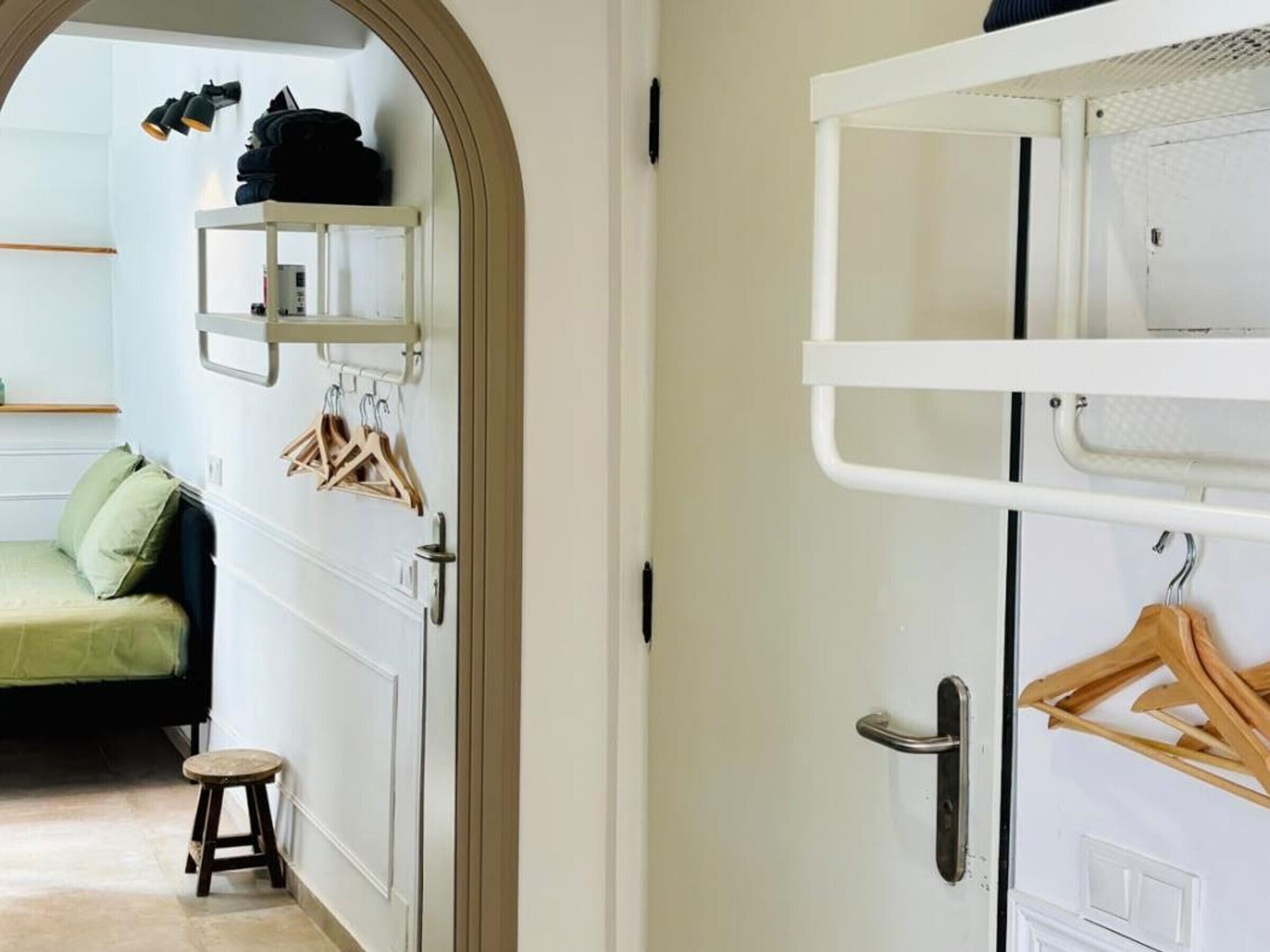 A room with a bunk bed, a small stool, a hanging rack with clothes, and a shelf with folded clothes.