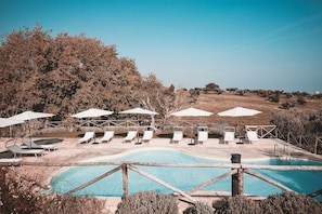 Un área de piscina con sillones de descanso y sombrillas, rodeada de árboles y un paisaje ondulado.