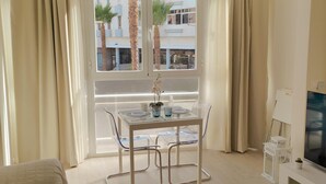Un salon moderne avec une table à manger en verre, des chaises transparentes, un canapé blanc, un téléviseur et une vue sur des palmiers et des immeubles, le tout visible à travers une grande fenêtre.