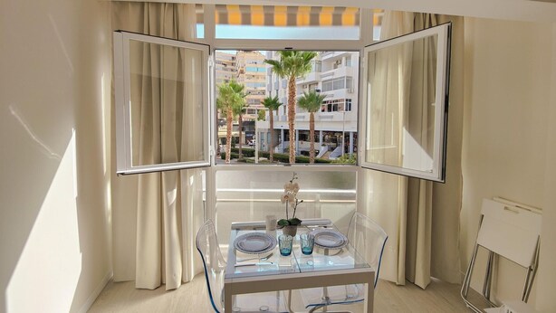 Une pièce moderne avec une table à manger en verre, des chaises transparentes et une vue sur les palmiers et les immeubles, le tout visible à travers des rideaux ouverts.