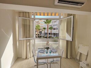 A modern room with a glass dining table, clear chairs, and a view of palm trees and buildings through open curtains. - Cozy Studio with Balcony in Torremolinos, Close to Beaches and City Center (Torremolinos)