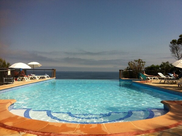 A pool with clear blue water, surrounded by lounge chairs and umbrellas, with a view of the ocean.
