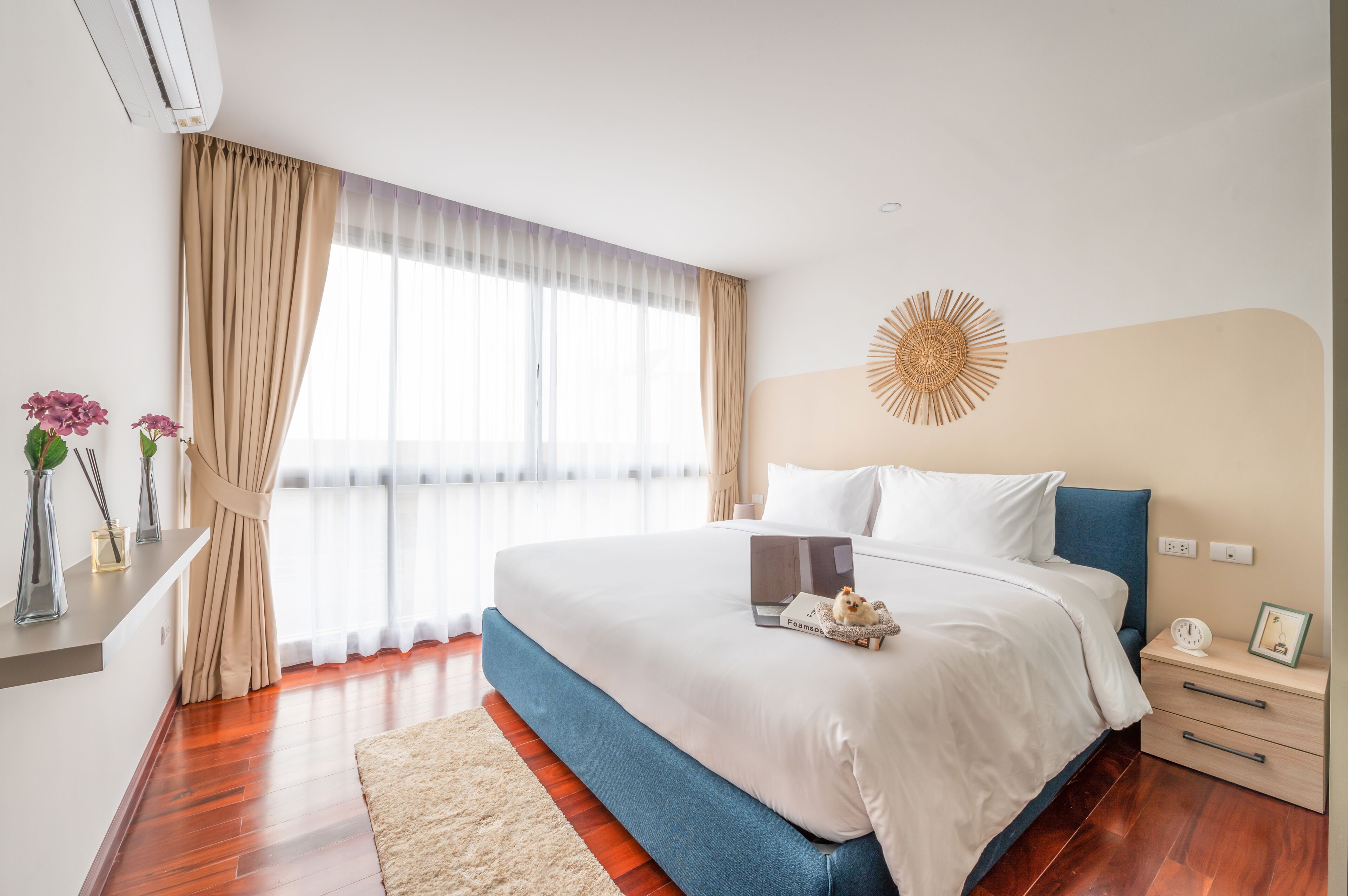 Una camera d'albergo moderna con un letto grande, un comodino con una lampada, un elemento decorativo a parete e una finestra con tende leggere.