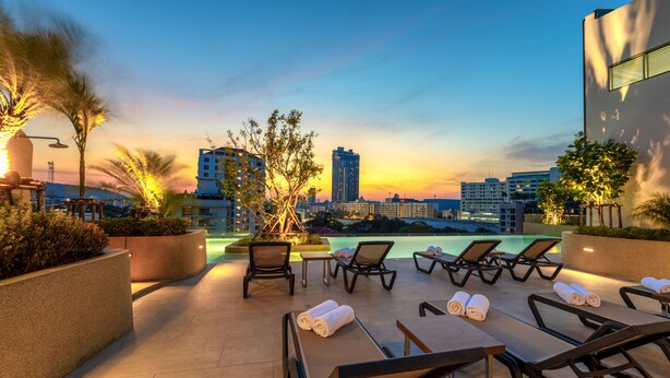 A rooftop lounge with sun loungers, a pool, and a city skyline view at sunset. - HOMA Si Racha (Si Racha)