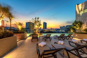 Un lounge en la azotea con sillones de sol, piscina y vistas a la ciudad al atardecer.
