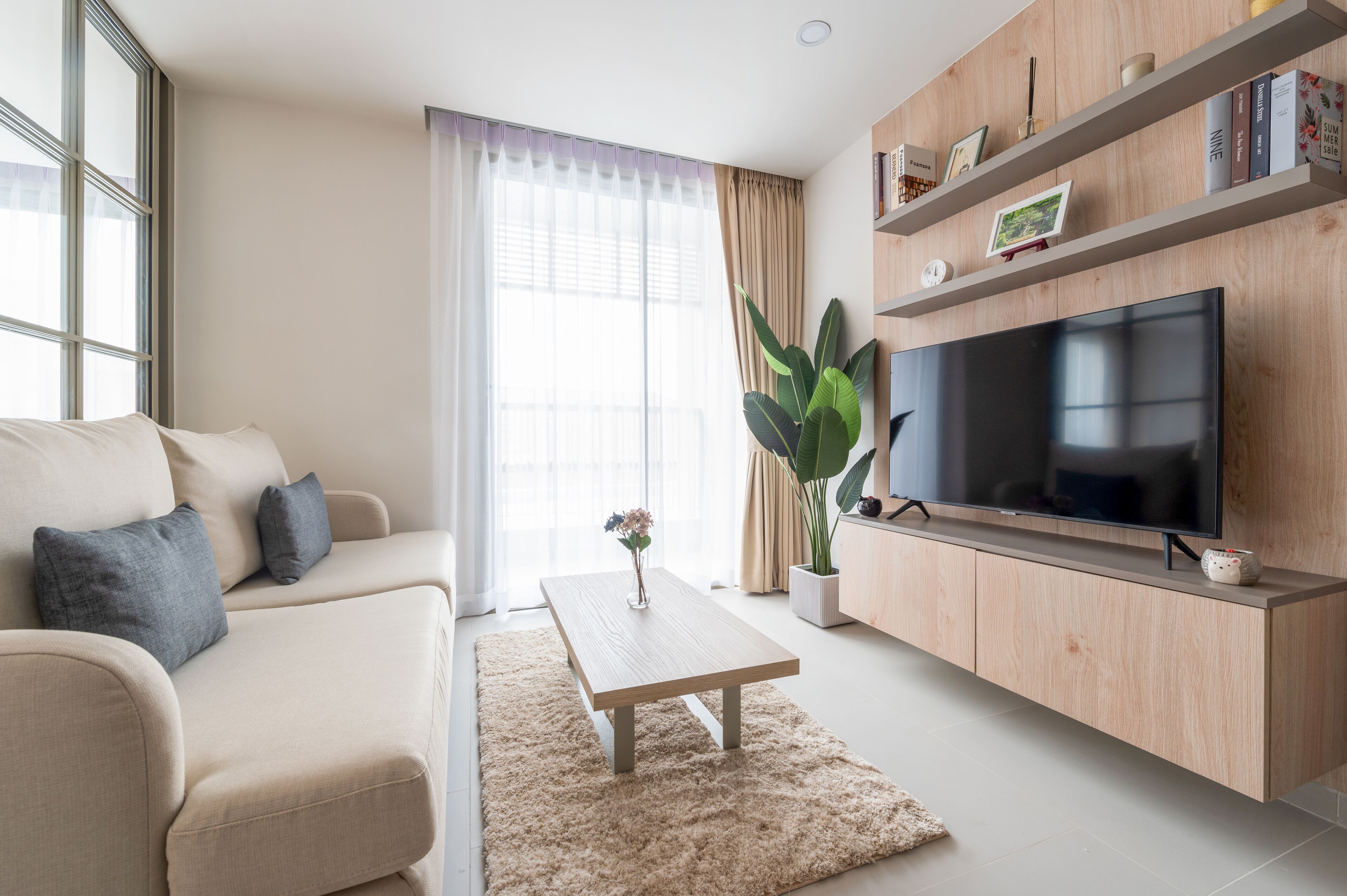 A modern living room with a beige sofa, a wooden coffee table, a large flat-screen TV, and a potted plant.
