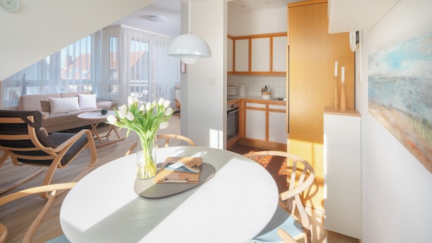 A modern living room with a round dining table, wooden chairs, a sofa, and a vase of white flowers. - // Dünensand Norderney // (Norderney)
