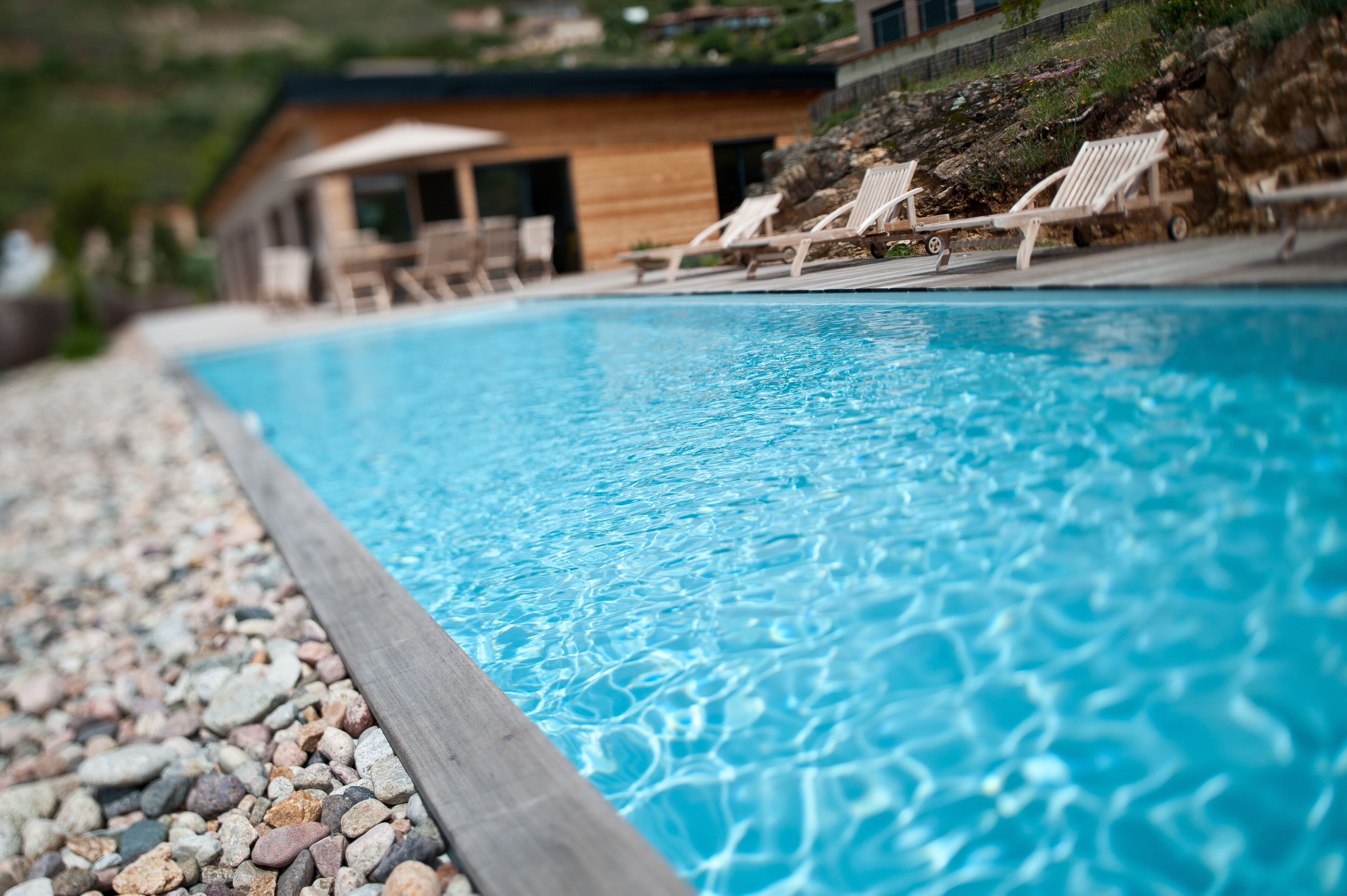A swimming pool with clear blue water, surrounded by a pebble border. There are lounge chairs and a wooden building with large windows in the background.