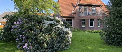 A two-story brick house with a red-tiled roof, surrounded by a well-maintained garden with flowering shrubs and a lawn.