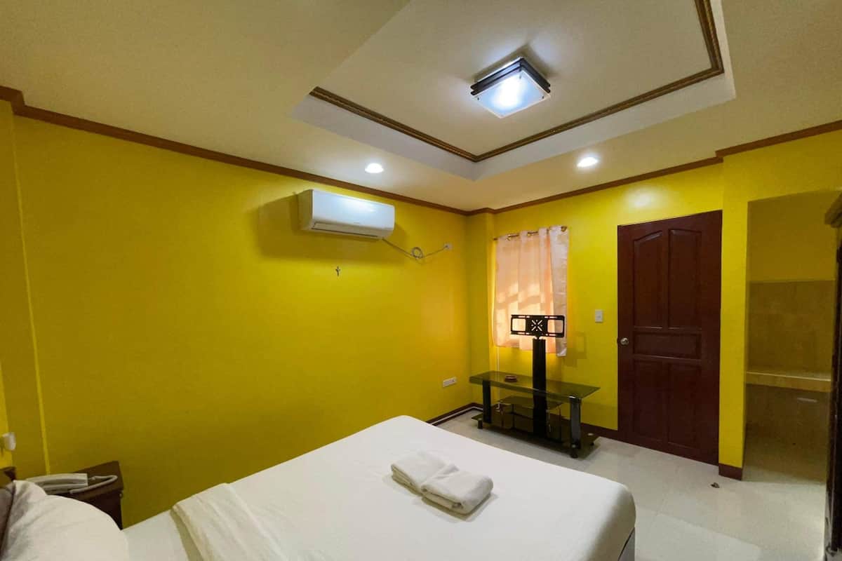 A hotel room with a bed, a small table, an air conditioning unit, a door, and a curtain.