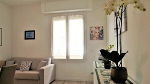 A living room with a beige sofa, a window with sheer curtains, a black vase with white flowers, and a framed picture on the wall. - Cristina Home "Town Center" (Sanremo)