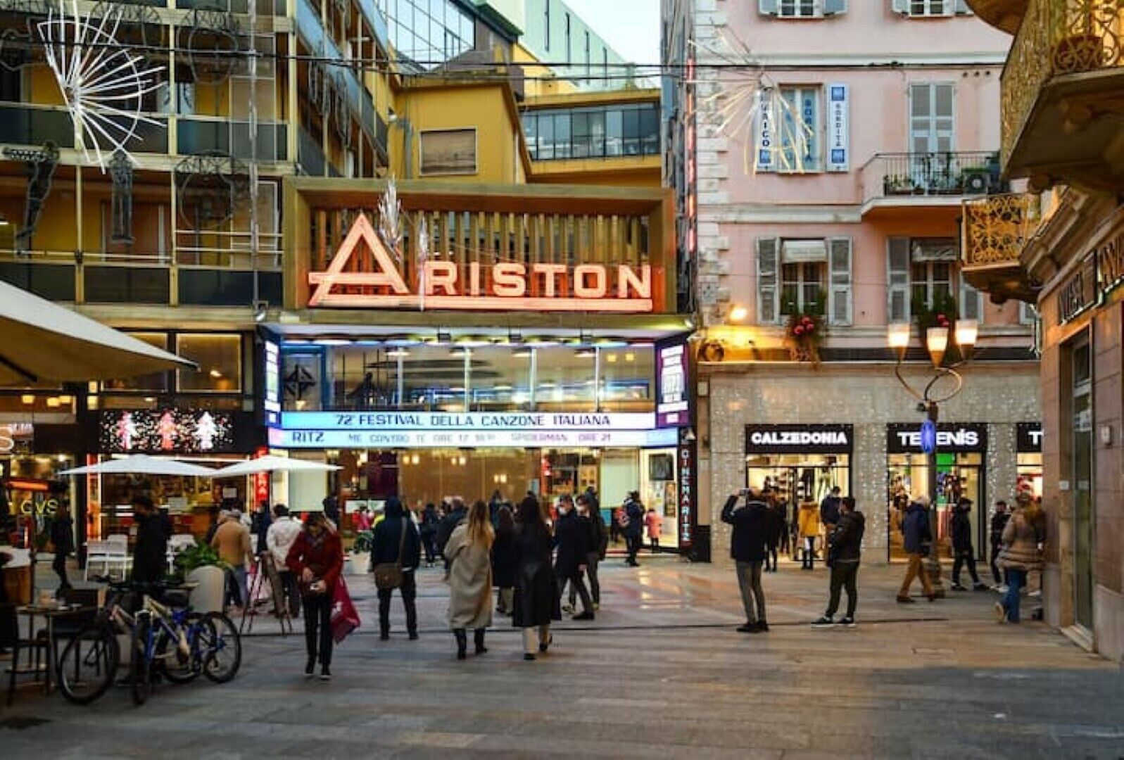 Una concurrida calle urbana con un edificio destacado que muestra un gran anuncio de 'ARISTON' y un cine.