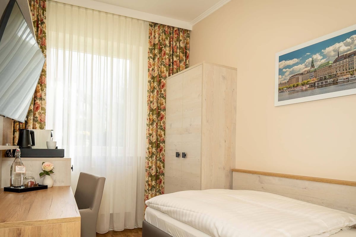 A hotel room with a bed, a desk with a chair, a wardrobe, and a painting of a cityscape.